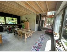 Offener Wohn- und Küchenbereich mit hoher Holzdecke und sichtbaren Balken, großem Esstisch, Küchenzeile und einem Holzofen. Rechts verlaufen bodentiefe Fenster entlang der Wand; im Hintergrund führen Stufen und eine Treppe zu einer verglasten Front mit Blick ins Grün.