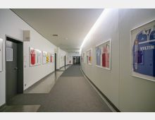 Ein langer, gewundener Korridor mit weißen Wänden, an denen gerahmte Fußballtrikots verschiedener Vereine hängen; der Boden ist teils mit Teppich, teils mit glattem Material bedeckt. Im Hintergrund sind Türen, Notausgangsschilder und ein Feuerlöscher sichtbar.