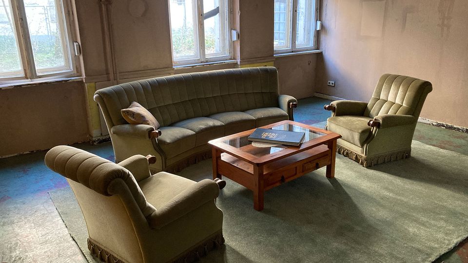 Karger, altmodisch eingerichteter Wohnraum mit großem Sofa und zwei Sesseln um einen Holz-Couchtisch auf einem grünen Teppich. Hohe Fenster lassen Tageslicht in den sonst spärlich möblierten Raum mit abgenutzten Wänden und Boden.