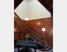 Treppenhaus mit dunkler Holzvertäfelung, einer dekorativen Holztreppe mit Balustern und Kugellampen als Beleuchtung. Im Vordergrund ein mit dunkelgrünem Tuch bedeckter Tisch, auf dem ein Blumenstrauß steht.