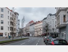 Städtische Straßenszene mit mehrstöckigen Wohngebäuden im klassischen Stil, teils mit Ladenfronten im Erdgeschoss. Am Straßenrand geparkte Autos, eine Fahrradfahrerin und Passanten sowie eine Kreuzung mit Verkehrsampeln sind sichtbar.