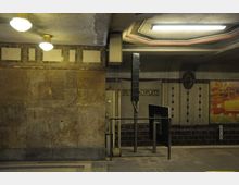 Untergrundbahnhof mit Vintage-Charakter, dunklen Fliesen und einer Beschilderung mit der Aufschrift „Breitenbachplatz“. Die Wände zeigen vergilbte Oberflächen, eine beleuchtete Decke und Wanddekorationen im Art-déco-Stil.