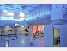 Innenraum eines wissenschaftlichen Labors mit einer modernen, futuristischen Einrichtung. Sichtbar ist eine große röhrenförmige Struktur mit dem Logo des DLR (Deutsches Zentrum für Luft- und Raumfahrt), ein Trainingsgerät mit kreisförmigem Rahmen sowie ein verglaster Raum mit Computer-Arbeitsplätzen und Stühlen.