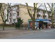 Ein kleiner Kiosk mit blauer Überdachung und Zeitungsständern steht am Rand einer Straße vor einem mehrstöckigen Wohnhaus mit hellgelber Fassade und Balkonen. Im Vordergrund stehen Bäume, und der Boden ist teils gepflastert und teils unbefestigt.