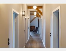 Flur mit Holzboden und weiß gestrichenen Wänden, gesäumt von Türen auf beiden Seiten. Sichtbare Holzbalken an der Decke und eine einfache Beleuchtung mit runden Lampen; am Ende des Flurs befinden sich ein Sessel und eine Kommode.