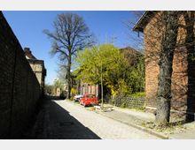 Gepflasterte Seitenstraße mit Ziegelgebäuden auf beiden Seiten, rechts flankiert von Bäumen und einem kleinen Zaun, links von einer hohen Backsteinmauer. Einige parkende Autos, darunter ein roter Wagen, stehen auf der rechten Seite, und der Himmel ist strahlend blau.