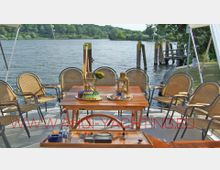 Eine Yacht-Terrasse mit einem gedeckten Holztisch, umgeben von geflochtenen Stühlen. Im Hintergrund ist ein ruhiges Gewässer mit grüner Uferlandschaft und einem kleinen Steg zu sehen.