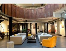 Moderner Lounge-Bereich in einem offenen Bürogebäude mit Sitzgruppen aus grauen Sofas und einem gelben Sessel, gläserner Konferenztür im Hintergrund, sichtbaren Rohren an der Decke und einer geschwungenen Wandverkleidung aus rostbraunem Metall. Links führt eine Treppe nach oben, rechts befinden sich Spinde und bunte Wanddekorationen.