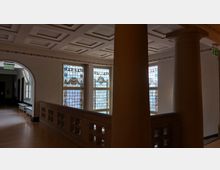 Flur in einem historischen Gebäude mit verzierten Buntglasfenstern, weißen Wänden und einem dekorativen Kassettendeckenmuster; im Vordergrund Säulen und ein Geländer mit rechteckigen Aussparungen.