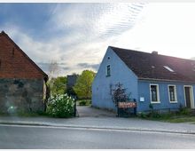 Ein ruhiger ländlicher Straßenzug mit einem blauen Gebäude rechts und einem Backsteinhaus links. Vor dem blauen Gebäude steht ein Schild mit der Aufschrift „Biergarten Do–Sa ab 12“, umgeben von Pflanzen und einem gepflasterten Weg im Hintergrund.