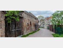 Eine schmale, gepflasterte Gasse mit einer alten Ziegelmauer und Holztüren auf der linken Seite. Rechts stehen Bäume und ein Zaun, während im Hintergrund mehrstöckige Wohngebäude zu sehen sind.