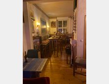 Schmaler, gemütlicher Gastraum mit Holzdielenboden, langen Holzbänken und hohen Tischen, an den Wänden große Bilder und warme Wandleuchten. Am Ende des Raums befindet sich ein Doppelfenster, daneben Regale und verschiedene Dekorationselemente.