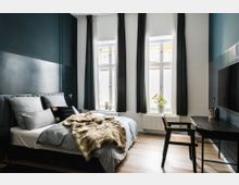 Modernes Schlafzimmer mit einem Doppelbett, dunkelblau gestrichenen Wänden und drei großen Fenstern mit Vorhängen. Im Raum befinden sich zudem ein Schreibtisch mit Stuhl, ein kleiner Fernseher und eine Zimmerpflanze auf der Fensterbank, der Boden besteht aus Holz.