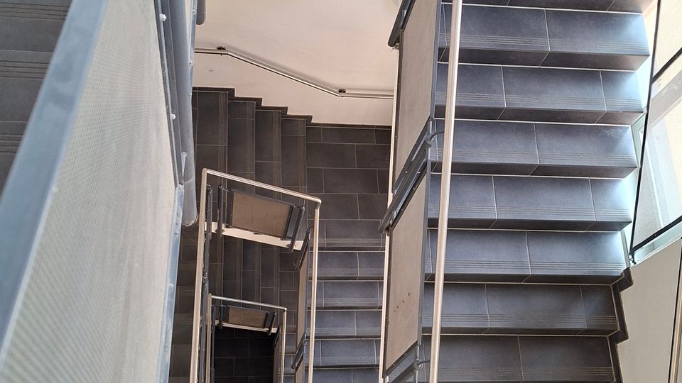 Blick von oben in ein modernes Treppenhaus mit mehreren Etagen, dunkel gefliesten Treppenstufen und Podesten. Metallgeländer mit Füllpaneelen rahmen den Treppenschacht, seitlich fällt Licht durch ein hohes Fenster.