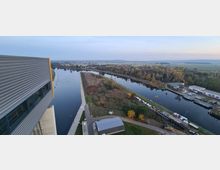 Blick von oben auf einen breiten Kanal, der von einer Grünfläche mit Bäumen und Sträuchern gesäumt ist, sowie auf ein modernes Gebäude mit grauer Metallverkleidung. Rechts sind ein Hafenbereich mit Booten und einige Industriegebäude zu sehen, umgeben von ländlicher Landschaft.