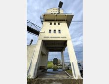 Ein alter Hafenspeicher mit der Aufschrift "Groß Neuendorf Hafen". Der Turm steht auf hohen Betonpfeilern, durch die ein Gleis mit einem historischen Güterwaggon führt; daneben befinden sich eine Wiese und parkende Fahrzeuge.