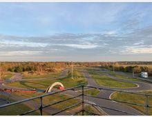 Go-Kart-Bahn in einer offenen, grünen Landschaft mit kurvenreichen asphaltierten Strecken, umgeben von Bäumen und Wiesen. Im Vordergrund ist ein Geländer zu sehen, und ein Bogen mit der Aufschrift „Go-Kartbahn.de“ markiert einen Teil der Strecke.
