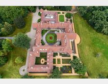 Luftaufnahme eines historischen Gebäudeensembles mit roten Ziegeldächern und einem Innenhof, in dessen Zentrum eine Rasenfläche mit einem roten Stern liegt. Umgeben ist die Anlage von gepflegten Grünflächen, Terrassen und Bäumen.