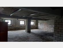 Ein leerer Kellerraum mit rauen Ziegelwänden, einem gemauerten Pfeiler in der Mitte und drei rechteckigen Fenstern, die Tageslicht hereinlassen. Der Boden besteht aus ungleichmäßig verlegten Ziegelsteinen, und die Decke ist niedrig mit sichtbaren Metallträgern.