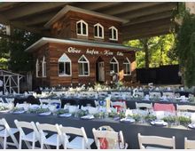 Eine rustikale Holzhütte mit der Aufschrift "Oberhafen-Kantine" steht unter einer Brücke, umgeben von Bäumen. Im Vordergrund sind lange Tische mit weißen Stühlen gedeckt, dekoriert mit Geschirr, Gläsern und Blumenschmuck.
