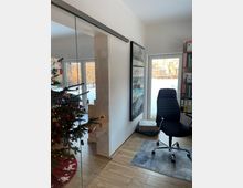 Heller Innenraum mit Schiebetür aus Glas, Holzoptik-Boden und Blick durch große Fenster und eine Glastür in einen verschneiten Garten. Rechts stehen ein schwarzer Bürostuhl auf einem Teppich und ein Regal, links ist ein geschmückter Weihnachtsbaum zu sehen.