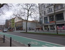 Straßenszene mit mehrspuriger Fahrbahn, Radwegmarkierungen in Grün und Fußgängern auf dem Gehweg. Auf der rechten Seite befinden sich Geschäftshäuser, darunter ein Edeka-Markt und eine Bankfiliale, umgeben von kahlen Bäumen.