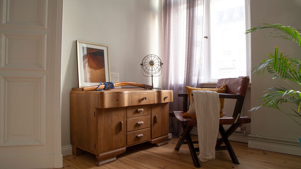 Heller Wohnraum mit Holzfußboden, großem Fenster mit transparenten Vorhängen und einem niedrigen Holzsideboard mit Dekoration. Daneben steht ein dunkler Stuhl mit Kissen und übergehängtem Tuch; eine Zimmerpflanze ragt am rechten Bildrand ins Bild.