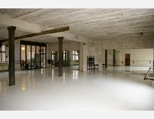 Großer Tanz- oder Übungsraum mit weißem, glänzendem Boden, alten Backsteinwänden und sichtbaren Deckenbalken. Die Ausstattung umfasst Spiegelwände, Stangen für Ballettübungen und große Fenster sowie Glastüren, die in einen angrenzenden Raum führen.
