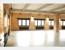 Offener Raum mit Sichtmauerwerk aus gelblichen Ziegelsteinen, schwarzen Heizkörpern und großen, rechteckigen Fenstern mit schwarzen Rahmen. Der Boden ist glatt und hell, die Decke hat freiliegende Betonbalken.