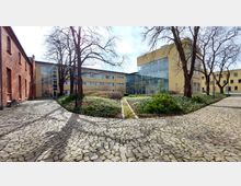 Gepflasterter Innenhof mit Beeten aus Grünpflanzen und Bäumen, umgeben von Gebäuden mit gelber Fassade und großen Glasflächen sowie einer roten Backsteinwand auf der linken Seite. Der Bereich wirkt gepflegt und zugänglich.