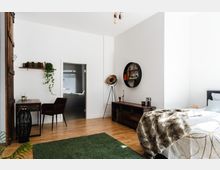 Schlafzimmer mit hellen Wänden und Holzboden, ausgestattet mit einem Bett, einem Schreibtisch mit Stuhl, einer Kommode und einer runden Wandregal-Dekoration. Ein grüner Teppich bedeckt einen Teil des Bodens, und eine Stehlampe sowie Zimmerpflanzen ergänzen die Einrichtung.