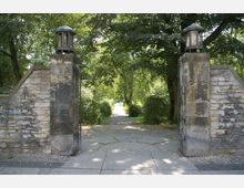 Steinerner Eingang mit zwei Säulen, die beidseitig mit kleinen, laternenartigen Aufbauten verziert sind, führt in einen grünen, von Bäumen und Sträuchern gesäumten Parkweg. Der Boden besteht aus großen, unregelmäßigen Steinplatten.