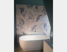 Modernes Badezimmer mit freistehender, ovaler Badewanne vor einer Wand mit einem großflächigen, dekorativen Design aus Koi-Fischen in Blau- und Grautönen. Auf dem Rand der Wanne stehen Pflegeprodukte, und der Raum hat eine minimalistische Gestaltung mit dunklen Wänden und Bodenfliesen.