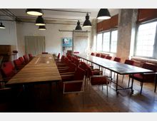 Ein Meetingraum mit langen Holztischen, umgeben von roten Stühlen mit Metallrahmen, auf einem Holzboden. Große Fenster mit braunen Jalousien lassen Tageslicht in den Raum, und rustikale Wände sowie Hängelampen aus Metall prägen das industrielle Design.