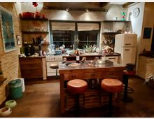 Gemütliche Küche mit gemauerten Wänden, einer zentralen Kücheninsel mit Holzoberfläche und Hockern mit orangefarbenen Polstern. Im Hintergrund befinden sich eine weiße Retro-Kühl-Gefrierkombination, ein Herd sowie offene Regale mit Gläsern, Flaschen und Küchenutensilien.