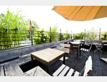 Holzterrasse mit einfachen Holztischen und -stühlen, umgeben von Pflanzen und einem Geländer mit hohen Gräsern. Ein großer Sonnenschirm spendet Schatten und schafft eine gemütliche, halboffene Atmosphäre.