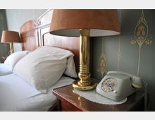 Ein Schlafzimmer mit einem Holzbettrahmen, weißen Kissen, einer Nachttischlampe mit goldenem Fuß und braunem Schirm sowie einem grauen Wählscheibentelefon auf einem Nachttisch mit Spitze. Die Tapete im Hintergrund hat ein florales Muster.