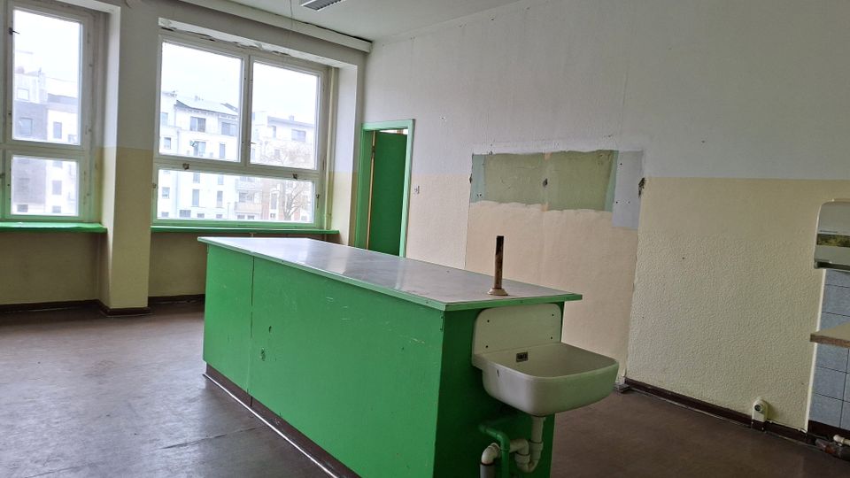 Leerer, schlicht eingerichteter Innenraum mit großen Fenstern und einem langen grünen Arbeitstisch mit integriertem Waschbecken und Wasseranschluss. Helle Wände mit abgenutzten Stellen und eine grün gestrichene Tür prägen den Raum.