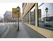 Blick auf einen städtischen Gehweg entlang eines gelben Gebäudes mit großen Fenstern, die Verkehrsschilder und eine Laterne spiegeln. Im Hintergrund sind Straßenkreuzungen, Verkehrsampeln und ein modernes, mehrstöckiges Gebäude sichtbar.