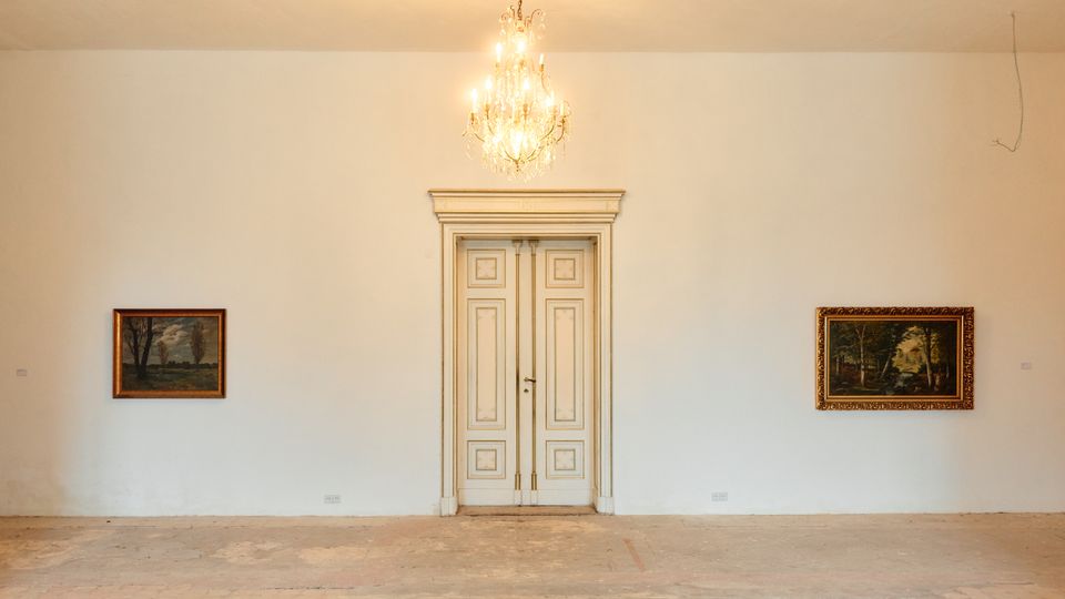 Heller Galerieraum mit weißer Wand, mittig einer hohen, klassisch gerahmten Doppeltür und darüber einem Kronleuchter. Links und rechts hängen gerahmte Landschaftsgemälde, der Boden ist roh und unbedeckt.