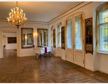 Elegant geschmückter Innenraum eines historischen Gebäudes mit Holzboden, großen Fenstern mit Vorhängen, reich verzierten Spiegeln und Wandgemälden. Ein Kronleuchter hängt von der Decke, und ein gedeckter Tisch mit rotem Läufer steht nahe einer doppelflügeligen Tür.