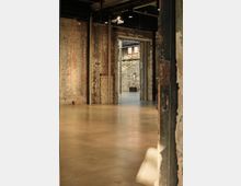 Industriehalle mit rohen Backsteinwänden und einem polierten Betonboden. Durch eine große Türöffnung ist ein benachbarter Raum mit ähnlichem industriellen Charakter zu sehen.