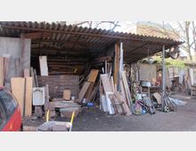 Überdachter Lagerbereich mit Wellblechdach, gefüllt mit alten Holzbrettern, Paletten und unterschiedlichen Gegenständen wie Fahrrädern, Werkzeugen und Schrottteilen. Die Umgebung ist ein Hof mit teils freiliegendem Boden, umgeben von Bäumen und kleineren Gebäuden.