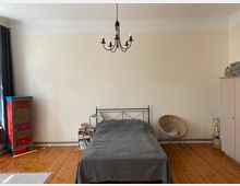 Ein schlicht eingerichtetes Schlafzimmer mit Holzboden, einem schwarzen Metallbett mit grauer Bettdecke in der Mitte, einer bunten Kommode links und einem geflochtenen Korbsessel rechts. An der Decke hängt ein schwarzer Kronleuchter, die Wände sind beige gestrichen.