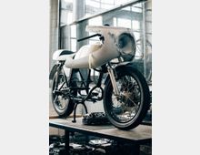 Ein unfertiges Motorrad mit rahmenlosem Verkleidungsaufbau steht auf einer Werkbank in einer Werkstatt mit großen Fenstern, durch die Tageslicht einfällt. Im Vordergrund sind Bauteile und Werkzeuge auf der Arbeitsplatte zu sehen.