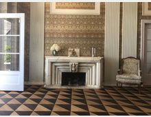 Klassischer Innenraum mit weißem Marmorkamin in der Mitte, gerahmt von hohen, geriffelten Wandpfeilern und einer gemusterten Tapete. Davor liegt ein schwarz-beiges Rautenkachelmuster am Boden; rechts steht ein gepolsterter Sessel, links ist eine geöffnete Glastür zu sehen.