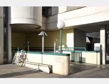 Moderner Außenbereich mit hellen Betonwänden, grünem Geländer und einer Rampe. Im Vordergrund steht ein Fahrrad an einer niedrigen Mauer mit Graffiti, daneben Straßenlaternen mit schrägen Metallabdeckungen.