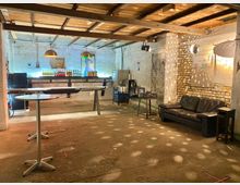 Innenraum einer rustikalen Bar oder Lounge mit unverputzten Wänden aus Ziegel- und Beton, einer langen Theke mit bunten Flaschen, einer schwarzen Ledercouch und einer Disco-Kugel, die Lichtpunkte an die Wände wirft. Die Decke besteht aus freiliegenden Holzbalken, und der Boden ist aus Beton.