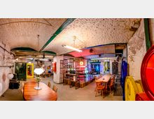 Großer, loftartiger Innenraum mit weiß gestrichenen Ziegelgewölbedecken, Sichtinstallationen und industrieller Beleuchtung. Lange Holztische mit Stühlen stehen vor einem abgetrennten Bühnenbereich aus Metallgittern; im Hintergrund sind farbige Neonlichter und ein Schild mit der Aufschrift „BERLIN“ zu sehen.