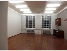 Ein leerer Raum mit Holzboden, weiß gestrichenen Wänden und zwei großen Fenstern mit Sprossen, die bis zur Decke reichen. Im rechten hinteren Bereich befindet sich eine kleine Küchenzeile mit dunklen Holzschränken, einem Herd und Fliesenrückwand.